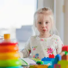 Mała dziewczynka z blond włosami w kwiecistym sweterku bawi się kolorowymi klockami.