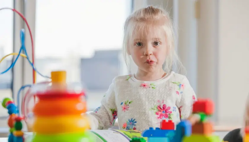 Mała dziewczynka z blond włosami w kwiecistym sweterku bawi się kolorowymi klockami.